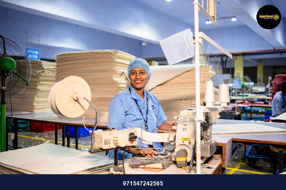Happy female textile worker in factory, operating a sewing machine with stacks of fabric.