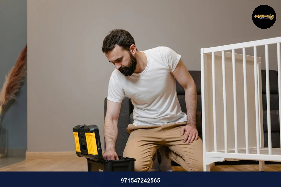 A man kneels indoors assembling a baby crib using a toolbox, wearing a white shirt and beige pants.