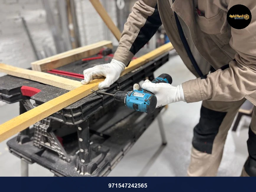 A carpenter using a power drill to work on wooden planks in an indoor workshop setting.