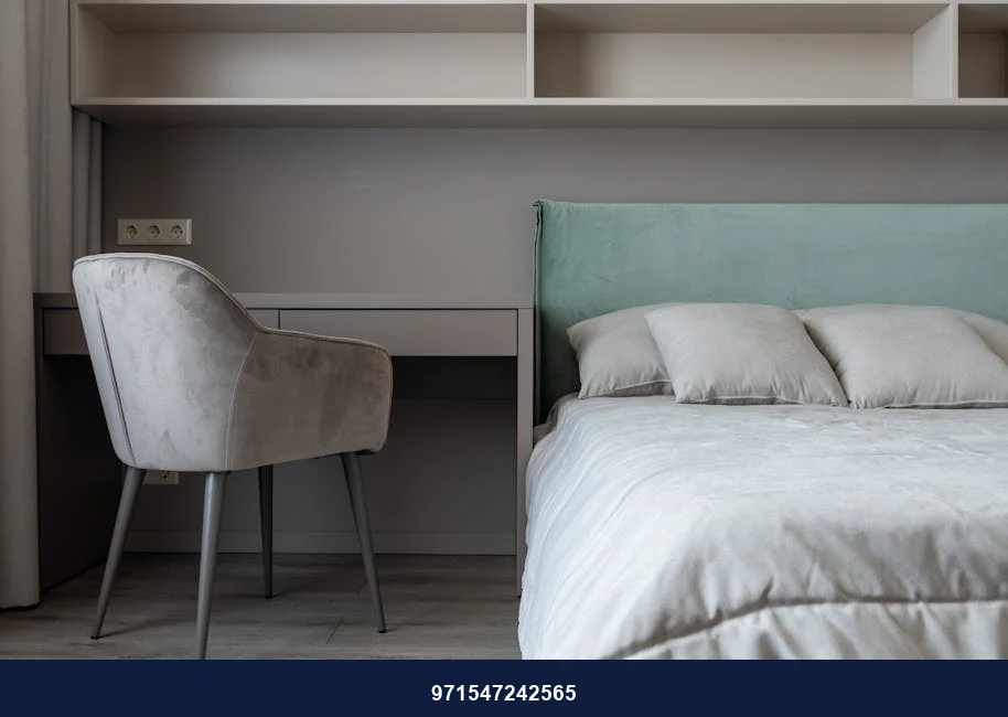 Interior of modern light bedroom with pillows and blanket on bed near table with chair under shelves next to curtains Interior of modern light bedroom with pillows and blanket on bed near table with chair under shelves next to curtains