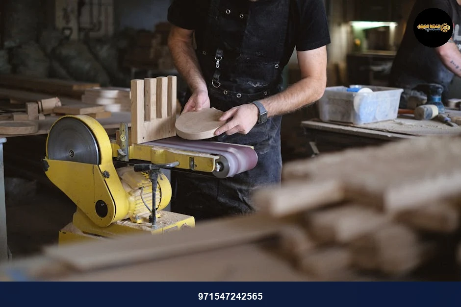 Unrecognizable male artisan using belt and disc sander to shape wooden detail while working near anonymous coworker in workshop with stack of wooden boards