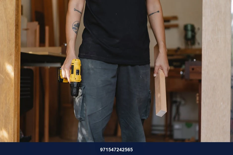 Unrecognizable craftsman in casual clothes standing with electric screwdriver and piece of wood while working in professional workshop