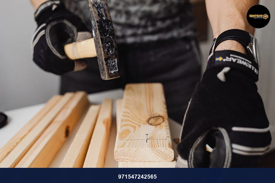 A carpenter wearing gloves uses a hammer to drive nails into wooden planks in a workshop.