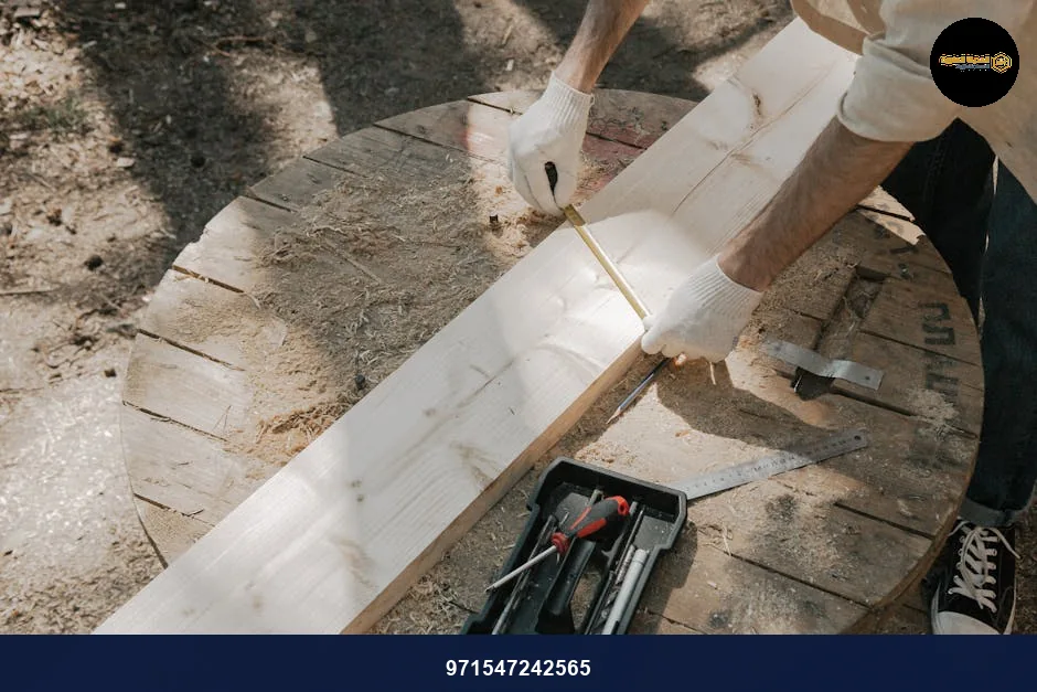 A carpenter in gloves measures a wooden plank on a round worktable with tools outdoors.