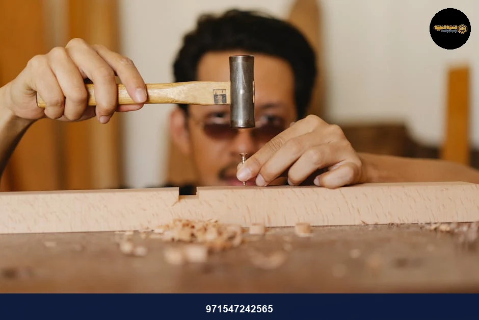 Concentrated young ethnic craftsman in eyeglasses hammering nail into wooden plank during work in carpentry studio