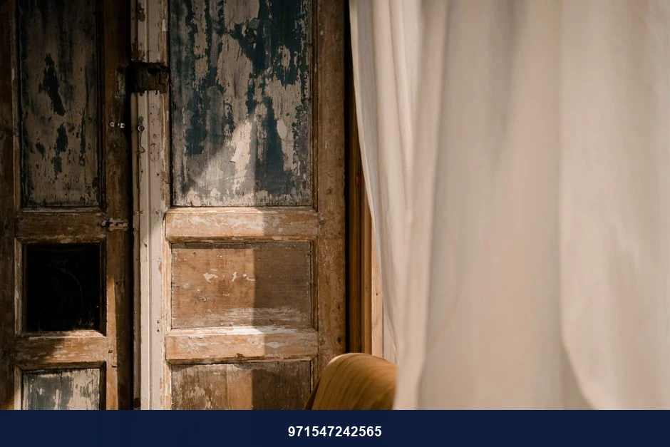 Sunlit rustic wooden door with aged texture partially covered by flowing curtains.
