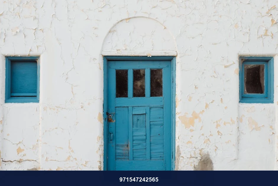 A quaint blue door on a weathered white wall with two blue windows.