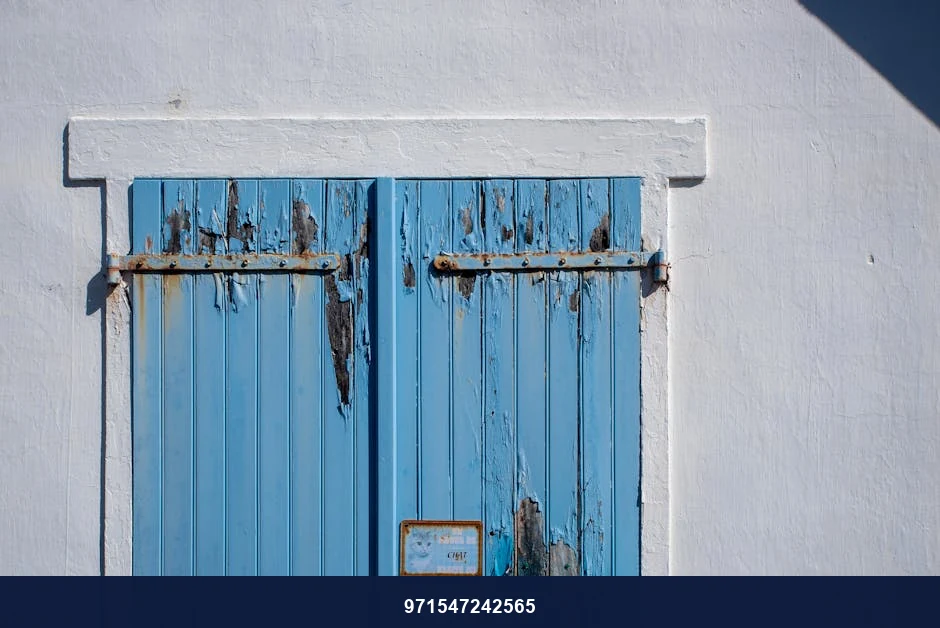 Old blue wooden shutters with peeling paint set on a white wall, displaying rustic charm.