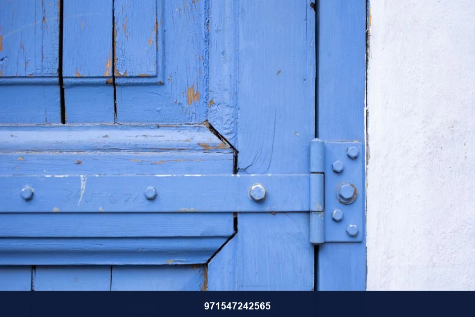 Fragment of weathered wooden door of blue color with metal hinge near white wall